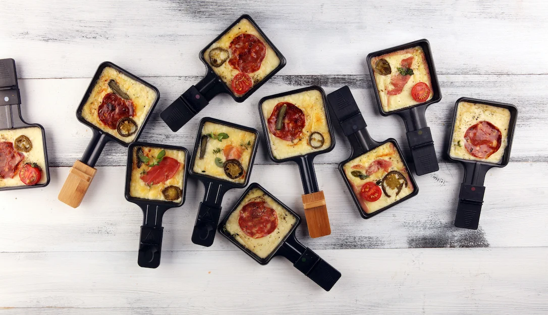 Gefüllte Raclette-Pfännchen stehen auf einem gedeckten Tisch.